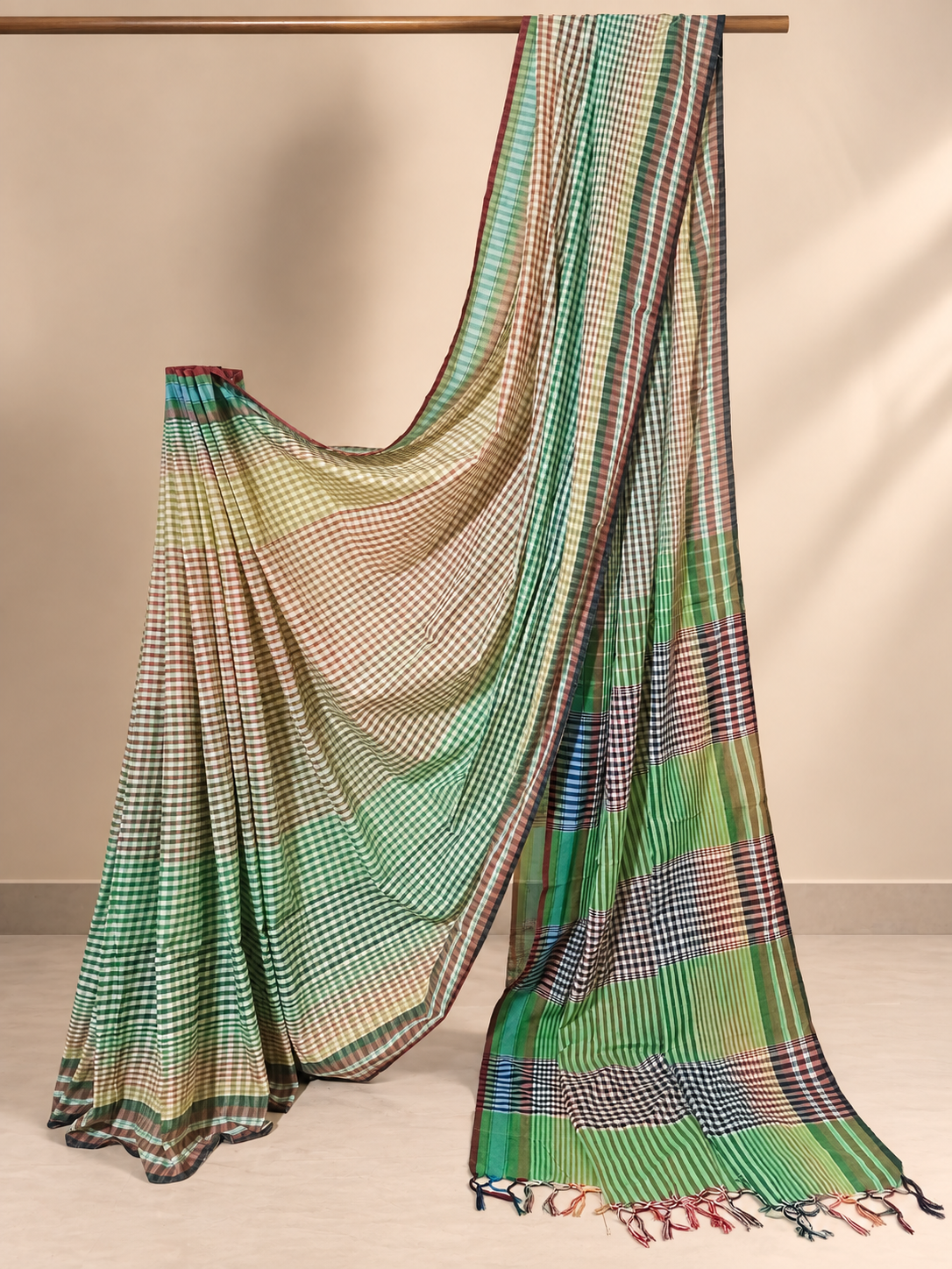 Green and cream handloom Gamchha saree with fine checked and striped patterns, an earthy multicoloured pallu and handcrafted tassels displayed in a minimalist studio setting.