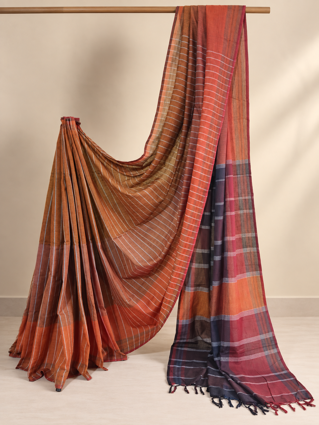 Rust and maroon handloom cotton saree featuring fine white stripes, contrasting checked sections and handcrafted tassels, displayed in a studio setting.